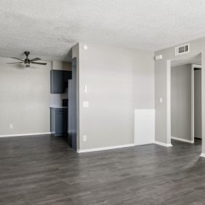 Empty living room with dark wood-style flooring, ceiling fan, and view into kitchen with blue cabinets.