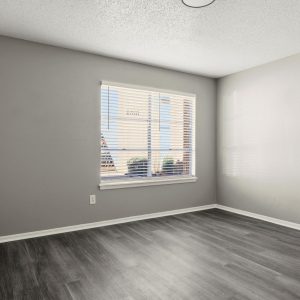 Empty bedroom with gray walls, dark wood-style flooring, and a large window with blinds overlooking city buildings.