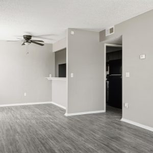 Empty renovated living area with gray plank flooring, ceiling fan, and pass-through opening to kitchen with black appliances.